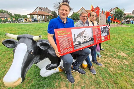 Bürgermeister Nicolas Aisch, Ortsvorsteher Werner Dürdoth und Pfarrer Werner Lütkefend präsentieren auf der symbolträchtigen  Kuh-Bank die Infotafel. Sie erläutert die Geschichte des Platzes und der Molkerei. Foto: Jürgen Vahle / Westfalen-Blatt 