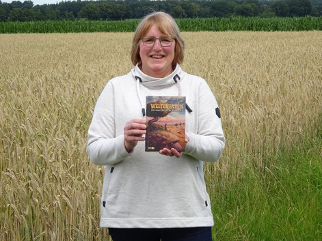 Ich stehe mit dem Buch "Weltenfäden" in der Hand auf dem Grasstreifen vor einem Getreidefeld. Ich trage ein helles Sweatshirt und Jeans.