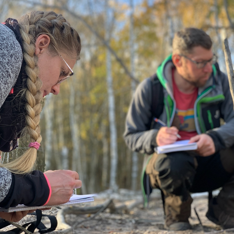 Zwei Teilnehmende machen kniend im Wald Notizen zu ihren Beobachtungen.