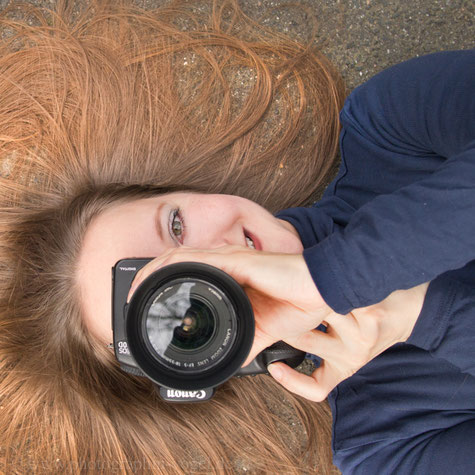 zu sehen ist ein Portrait von Ute Vogel-Rusche, Inhaberin vom Photostudio NEUES LEBEN  auf Asphalt liegend mit ausgebreiteten langen blonden Haaren und in Photograhierpose mit der Kamera vor dem halben Gesicht