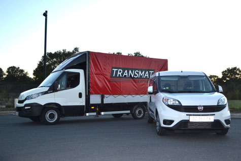 Un camion bâché rouge de la société Transmat et un petit véhicule utilitaire blanc garés côte à côte sur une route, prêts pour des services de transport ou de livraison.