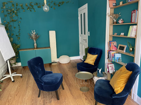Ein Coachingraum mit 3 sesseln, 2 tischen und einer türkiesen wand. Bücher und deko sind auch zu sehen