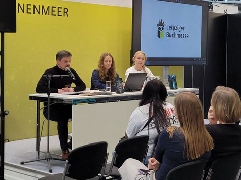 Drei Personen sitzen nebeneinander und lesen aus Büchern vor einem Publikum.