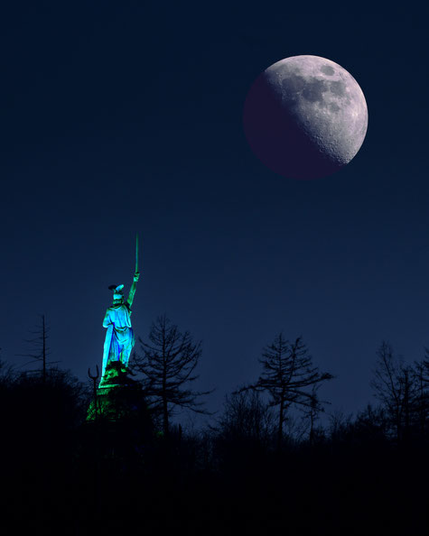Hermann reckt sein Schwert gegen den aufgehenden Mond – das nächtlich beleuchtete Denkmal erscheint wie ein mythisches Sinnbild von Sieg und Freiheit, eingerahmt von dunklen Baumwipfeln vor nachtblauem Himmel