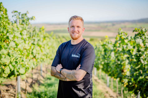 Mann in schwarzem Woihesse Unisex-Shirt mit weißer Stickerei, steht lächelnd im Weinberg in Rheinhessen – nachhaltige Mode mit regionaler Botschaft von Rhoihesse on Tour.