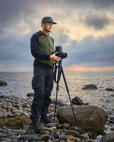 Fotograf Christopher Rau steht mit einer Analogen Mittelformatkamera auf einem Stativ an einem steinigen Strand der Insel Rügen, im Hintergrund die abendlich beschienene Ostsee.