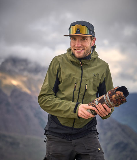 Fotograf Christopher Rau trägt grüne Outdoorjacke und schwarze Hose, hält eine getarnte Kamera mit großem Teleobjektiv, vor Berglandschaft bei bewölktem Himmel