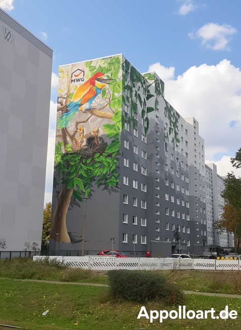 Fassadenbild eines bunten Bienenfressers auf einem Ast über einem Nest mit drei Jungvögeln, gemalt an der Seite eines hohen, grauen Wohngebäudes der MWG. Der Himmel ist bewölkt, und Stromleitungen sind im Vordergrund sichtbar.