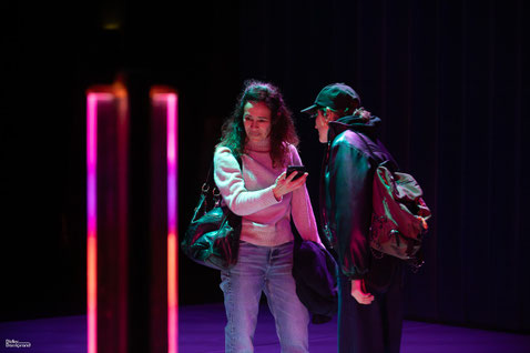 Photo du spectacle M'aime pas mort. La journaliste fait face à Gone, elle l'enregistre avec son téléphone portable. Gone porte une casquette. Ambiance lumineuse particulière avec des néons roses et bleus.