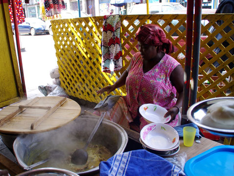 Afrikanerin in Straßenküche in Togo hinter großem Kochtopf und Geschirr sitzend