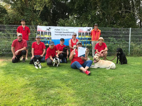 Gruppe von Personen in roten Shirts mit mehreren Hunden vor einem Zaun mit Bannern der Flyball-Mannschaft Tübinger Hundefreunde Hard Fighting Devils auf Gras.