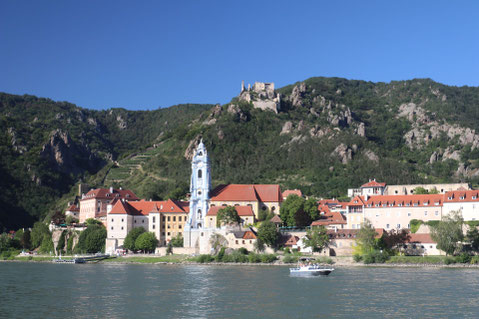 Dürnstein in der Wachau vom gegenüberliegenden Ufer der Donau aus mit markantem blauem Kirchturm und grün bewachsenen Felsen in Hintergrund