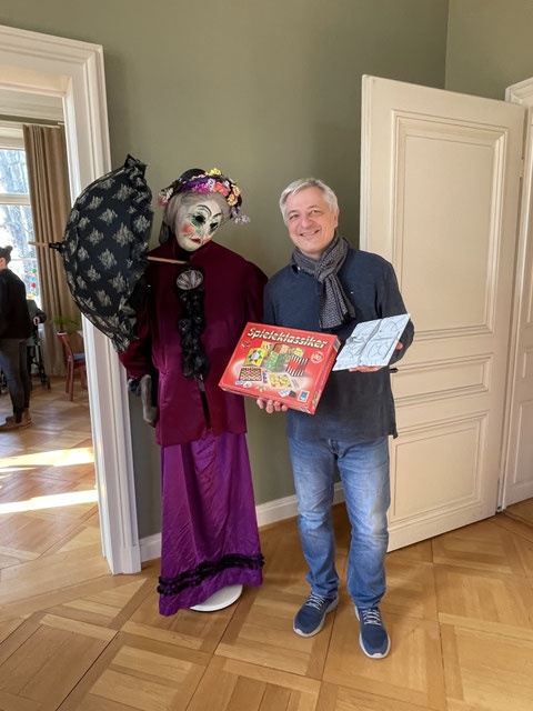 Christian Gilgen neben der Figur einer faschnächtlichen Alten Tante; er hält eine Schachtel mit Spielen und ein Geschenk in der Hand