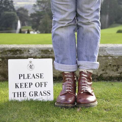 Person mit Schuhen auf Gras, daneben ein Schild: Please Keep Off The Grass