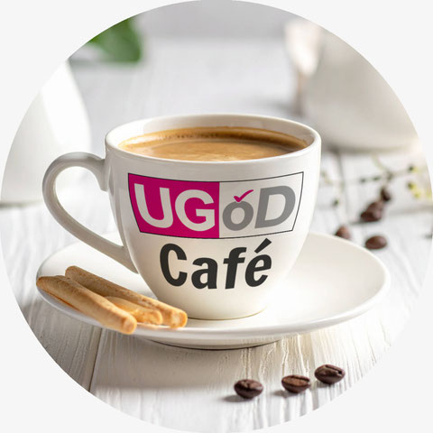 Eine Tasse mit Kaffee steht auf einem Tisch. Auf der Tasse ist aufgedruckt: UGÖD-Café