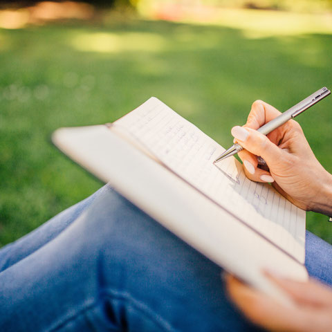 Eine Studentin sitzt im Gras und schreibt in ihrem Heft