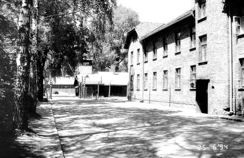 Blick aus dem Lager auf das Lagertor mit Schlagbaum und Blockführerstube im Hintergrund. Im Block 24 (sog. Schreibblock, rechts) war die Häftlingsschreibstube untergebracht.