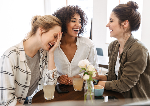 Drei junge Frauen sitzen fröhlich lachend an einem Tisch.