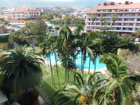 Hinter subtropischen Pflanzen der Gartenanlage sieht man den Pool und am Horizont den Teide. 