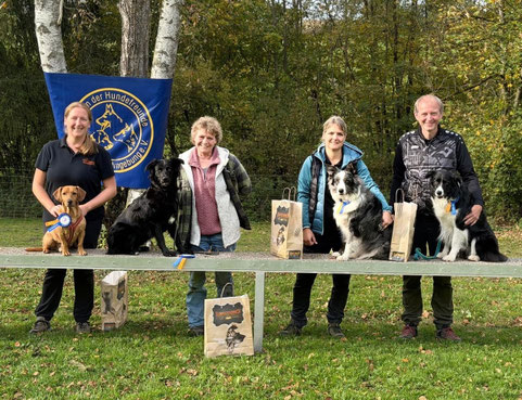 Vier Personen mit jeweils einem Hund unterschiedlicher Rassen stehen hinter einer Bank im Freien, auf der mehrere Papiertüten mit Hundemotiven stehen.