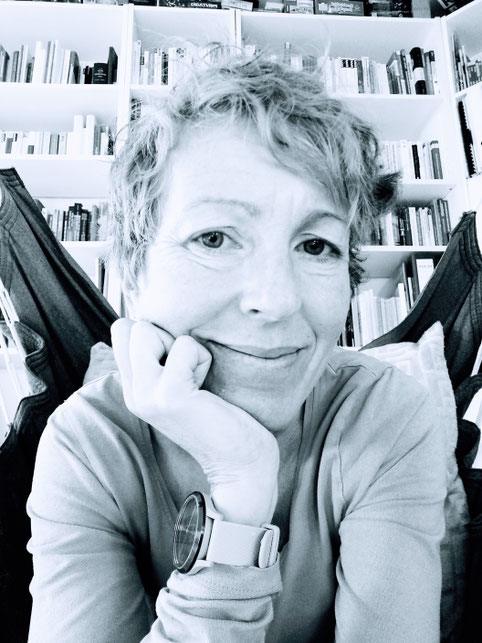 Schwarz-weißes Porträt von Christine Harnik mit kurzem Haar, die den Kopf auf die Hand stützt und leicht lächelt; im Hintergrund sind Bücherregale zu sehen.