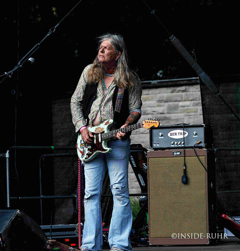 <img src="zen-trip-live.jpg" alt="Gitarrist der Band Zen Trip spielt eine weiße Fender Stratocaster mit rotem Gurt auf einer Bühne. Er trägt ein gemustertes Hemd und blaue Jeans, hat langes Haar. Neben ihm steht ein Verstärker mit der Aufschrift 'Zen TRIP