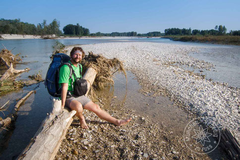 Wanderer sitzt gemütlich auf einem Baumstamm und streckt die Füße in die Piave