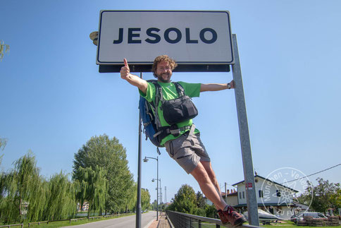 Wanderer umarmt glücklich das Ortsschild von Jesolo und hält dabei den Daumen hoch