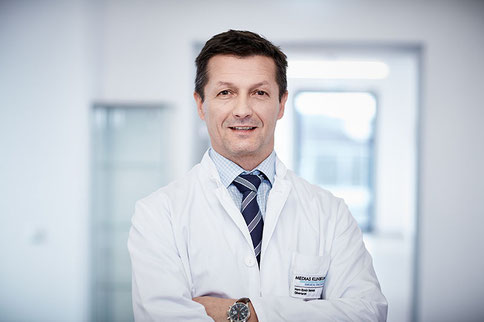 Herr Emir Selak, Leitender Oberarzt, im weißen Kittel, steht in einem modernen Klinikflur und lächelt freundlich in die Kamera.
