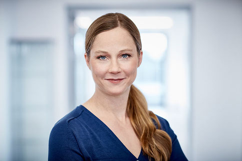 Kornelia Aigner, Dipl.-Biologin, im blauen Oberteil, steht in einem modernen Klinikambiente und lächelt freundlich in die Kamera.