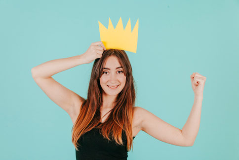 A smiling woman wearing a paper crown, raising her arm in triumph, symbolizing success and joy in learning Spanish.