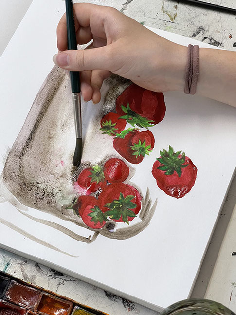 Zu sehen ist die Hand einer jungen Frau, die mit Aquarellfarben ein Stillleben mit Erdbeeren malt