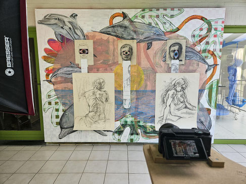 Groot samengesteld werk met tekeningen, dolfijnen en figuren tegen een wand; op de voorgrond een camera op een houten steun die de opstelling registreert.