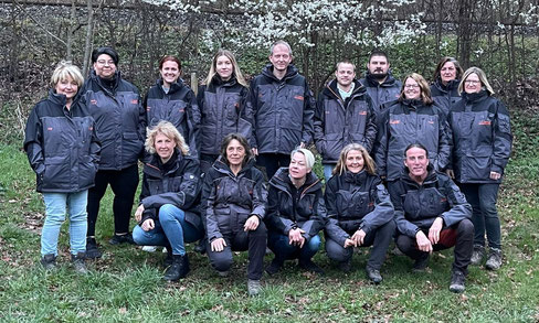 Gruppe von Personen in dunklen Jacken vor blühendem Baum im Freien, einige stehen, andere knien auf Grasboden.
