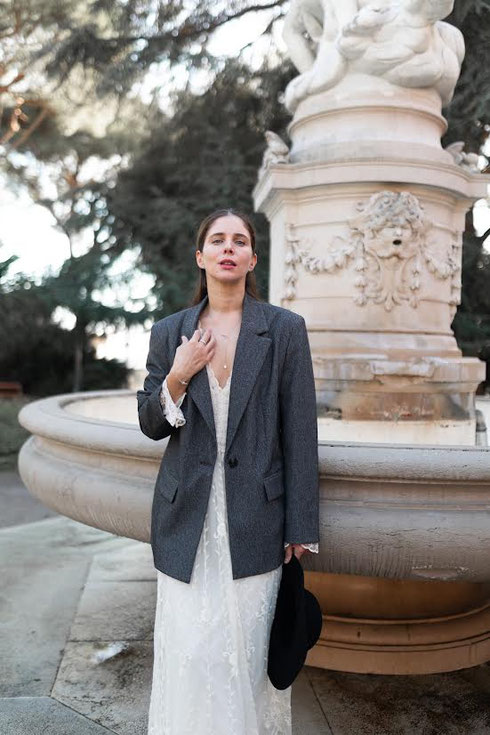 Woman in a white lace dress and grey blazer standing in front of an ornate stone fountain. She holds a black hat, creating a calm and stylish atmosphere.