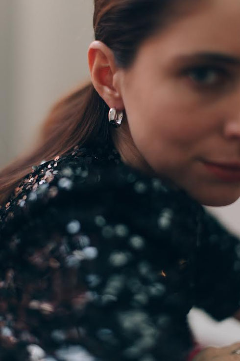 Portrait of a woman in black shimmering clothing wearing silver hoop earrings. The focus is on the elegant earring while the rest of the image is softly blurred.