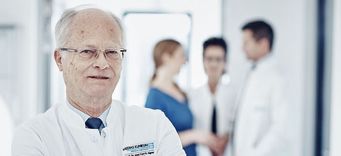 Arzt im weißen Kittel steht im Vordergrund, während im Hintergrund zwei weitere Ärzte mit einer Patientin im Gespräch sind.