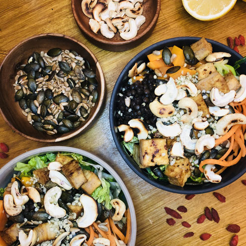Zwei Tonschalen, grau und beige, bunt gefüllt mit Tofu, Cashews, Belugalinsen, Süßkartoffel und Karotte. Daneben stehen in Holzschälchen Kürbiskerne und Sonnenblumenkerne. Rechts oben liegt eine halbe Zitrone und um die Schalen herum rote Gojibeeren.