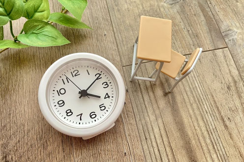 目覚まし時計と小さな机・椅子のミニチュア。忙しい社会人が勉強時間を確保するイメージ。