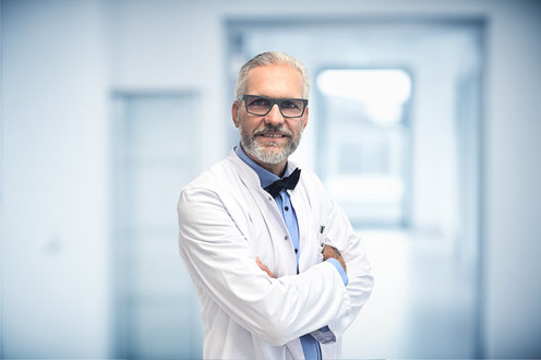 Portrait eines freundlichen Arztes im weißen Kittel mit Brille und grauem Haar in modernem Klinikflur. PD Dr. med. Hansjörg Aust blickt selbstbewusst mit verschränkten Armen in die Kamera.