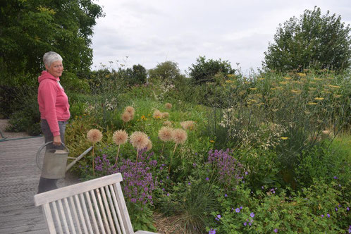 Eine Frau mit einer Gießkanne in der Hand steht auf einer Terrasse vor einem sommerlichen Staudenbild. Der Hintergrund zeigt Bäume und einen bewölkten Himmel.