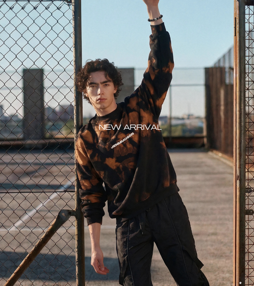 men model standing on the doorway of a metal grid fence wall,  wearing black techwear cargo pants with a black and orange acid-washed sweater with white letter print on the front