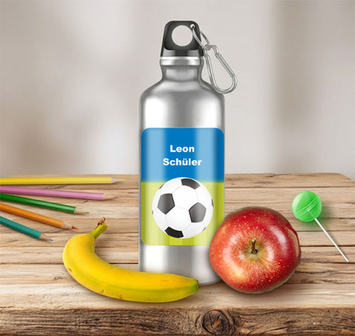 Silberne Trinkflasche mit Fußball-Namensaufkleber auf einem rustikalen Holztisch neben Obst, Stiften und Lolly.