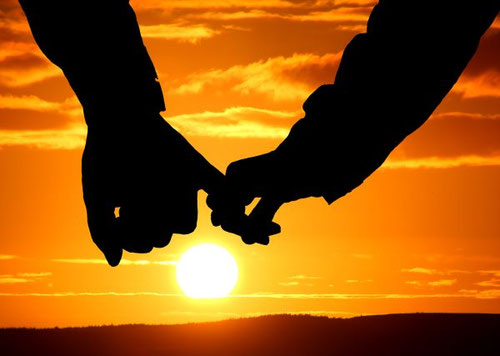 Zwei Hände von zwei Personen halten sich vor einem orangefarbenen Sonnenuntergang.