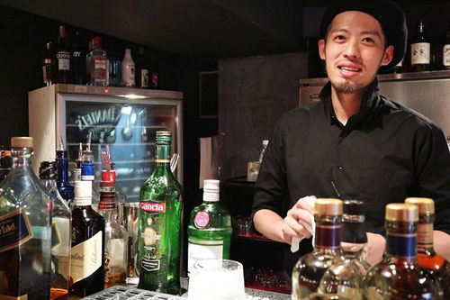 カウンターに立つ沓沢店長の写真。カウンターに並ぶ様々な酒瓶。