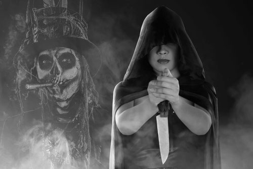 voodoo ritual frau mit kaputze auf dem kopf und messer in der hand steht vor flammen mit baron samedi im hintergrund