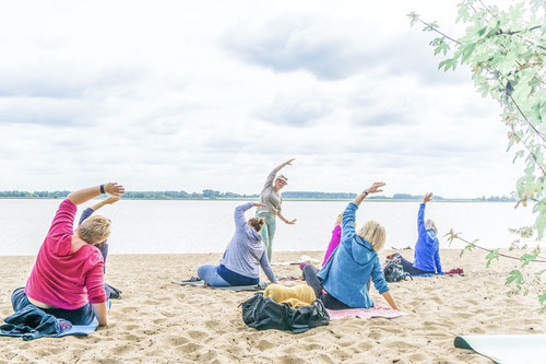 Eine Gruppe praktiziert Yoga am Elbstrand mit Blick aufs Wasser unter Anleitung von Yogalehrerin Rieke Buning