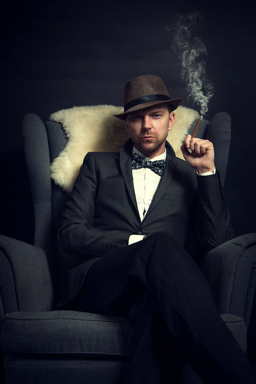 A man in a suit and fedora sits confidently in a chair, holding a smoking cigar with a serious expression against a dark background.