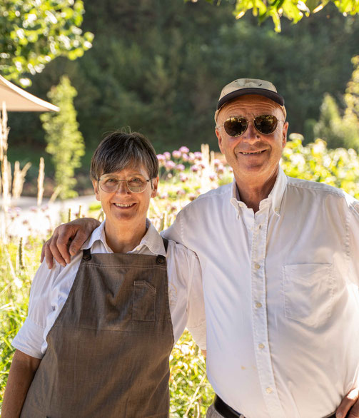 Zwei lächelnde Personen stehen Arm in Arm in einem sonnigen Garten; die Frau trägt eine Schürze, der Mann eine Kappe und Sonnenbrille.