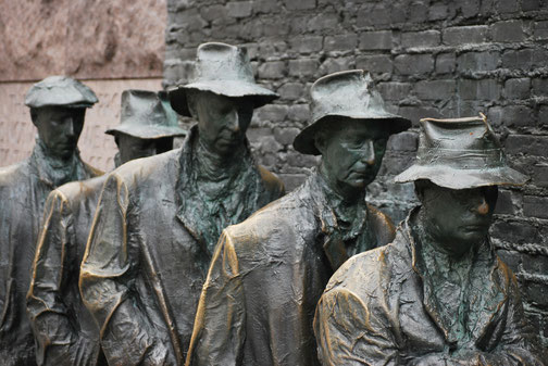 Bronze statues of weary men in hats stand in a line against a brick wall, representing a breadline during the Great Depression.